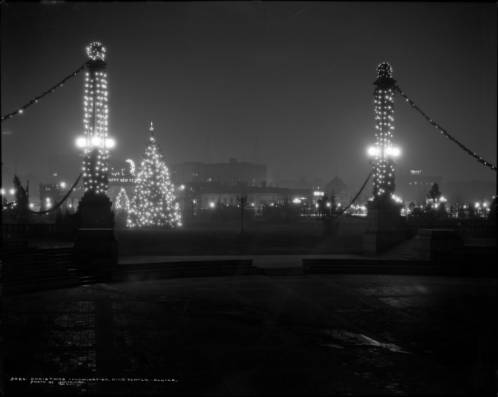 View northeast from Greek Theater and Colonnade of Civic Benefactors across Civic Center Park to Voorhies Memorial and downtown Denver, Colorado; illuminated Christmas decorations include: large Christmas tree center of plaza, evergreen wrapped lampposts and columns, "Happy New Year" on Voorhies Memorial; landmarks include: Continental Oil Building with large illuminated sign, "Conoco Products," Metropolitan Building, Majestic Building, Western Tire Co., and YMCA. (1929)
